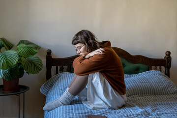 Apathetical unemotional introverted teenager sitting on bed embracing knees alone at home....