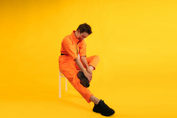 Middle age funny and Cheerful woman wear modern Orange overalls sitting on white chair over yellow color background. Female having fun and energetic movement studio shot 