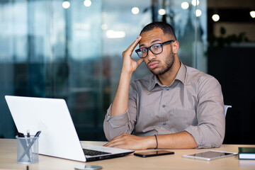 Frustrated and sad man at workplace reading online message from laptop, businessman sitting inside office at workplace, desperate unhappy, got bad news, bankruptcy, layoff. - obrazy, fototapety, plakaty