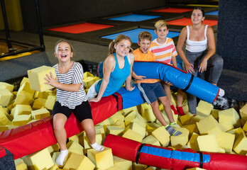 Company of joyful smiling children sitting together on crossbar in entertainment trampoline centre...