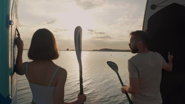 Happy couple preparation for paddle boarding at lake during sunset together Back view. Healthy people enjoy outdoor active lifestyle play extreme sport surfing on summer vacation.