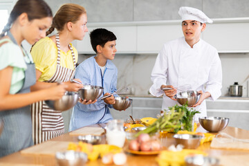 Young guy cook in uniform teaches group of children how to cook dish