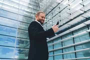 Fototapeta premium Cheerful businessman using smartphone near glass building