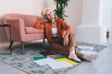 Cheerful mature woman sitting on floor with book and papers in living room