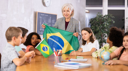 Mature female high school teacher, conducting a lesson in the classroom, holds the national flag of Brazil and tells the pupils .the history of the country