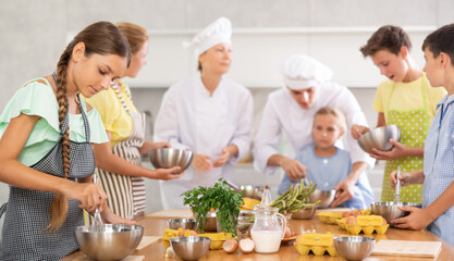 During cooking classes, girl carefully mixes ingredients in bowl with whisk. In background, blurry children stand near kitchen table and listen to chefs explanations