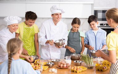 At culinary master class, experienced male chef in white uniform explains to children subtleties of dessert preparation and female assistant helps child to work with whisk