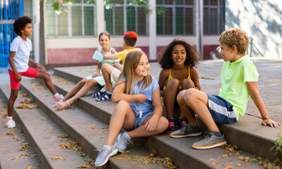 Happy tweenagers friends spending time together in city streets during summer vacations, sitting on...