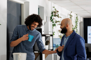 Corporate colleagues telling joke and laughing while drinking coffee in coworking space. Smiling young arab start up company employees enjoying work break in business office - obrazy, fototapety, plakaty