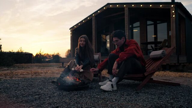 A couple of friends guy and girl sitting together around a campfire in woods making marshmallows and chatting animatedly