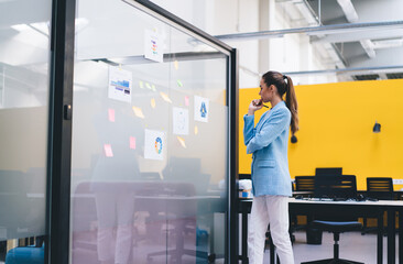 Young pensive woman standing by glass wall with sticky notes in office