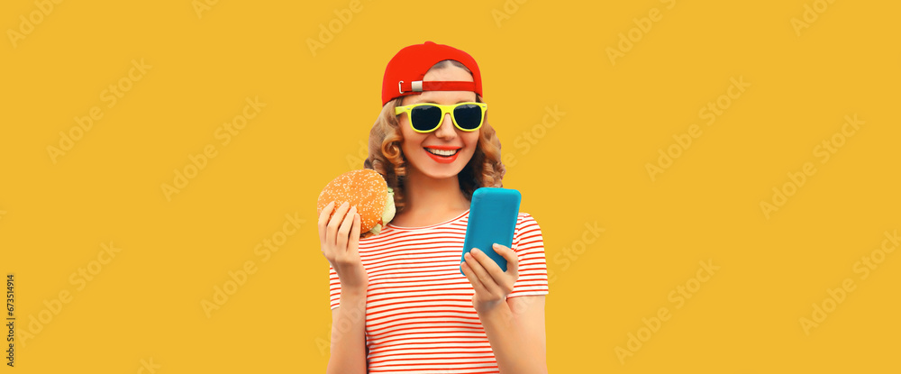 Wall mural portrait of stylish happy smiling young woman with mobile phone and burger fast food on yellow studi