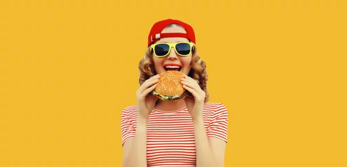 Poster Portrait of stylish happy smiling young woman eating burger fast food on yellow studio background © rohappy
