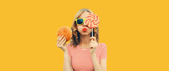 Portrait of cheerful young woman with burger fast food and colorful lollipop on yellow studio background
