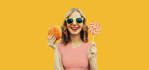 Portrait of cheerful young woman with burger fast food and colorful lollipop on yellow studio...