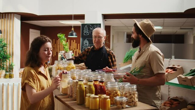 Elderly vendor assisting clients in eco friendly shop with ethical sourced food items, providing recommendations. Woman and man in local store chatting with trader about bulk products