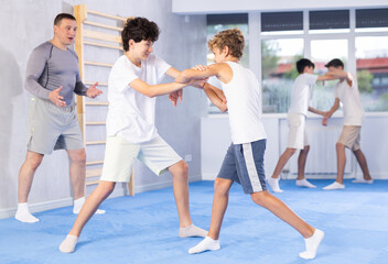 Fototapeta premium Focused teenage boys practicing close combat techniques in sparring in training room during self-defence workout under supervision of coach