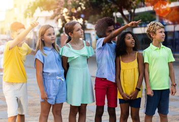 Group of happy inquiring preteen friends of different nationalities looking with interest at something and pointing with hand while walking on summer city street