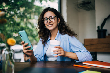 Cheerful woman with smartphone and coffee sitting at table in cafe