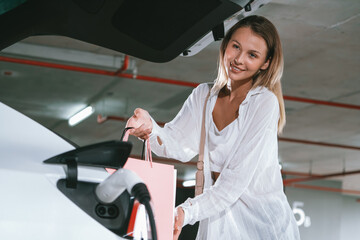 Young woman travel with EV electric car to shopping center parking lot charging in downtown city...