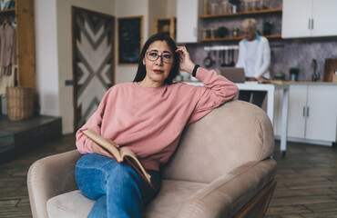 Obraz premium Cheerful middle aged ethnic woman sitting with book on sofa