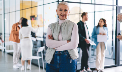 Positive senior woman with crossed arms against blurred colleagues in office
