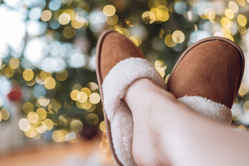 Christmas and New Year holidays.legs in brown suede fur slippers on a Christmas tree in garlands background.Winter comfort and holidays. Festive mood.