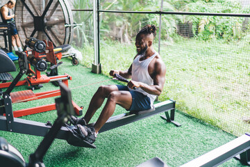 Cheerful black sportsman using pulling strong cable on bench in gym