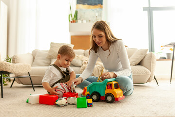 mother with her little son sits on the carpet at home and plays with toys in the room, female teacher with boy child - obrazy, fototapety, plakaty