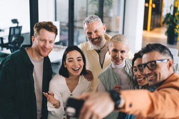 Obraz premium Cheerful male and female colleagues enjoying friendly cooperation making selfie images on smartphone in office interior, happy group of entrepreneurs clicking cellular photos and smiling at camera