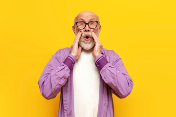 shocked old bald grandfather in glasses looks in surprise at the camera on a yellow isolated...