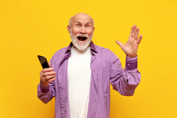 surprised old bald grandfather uses a smartphone and screams with joy on a yellow isolated...