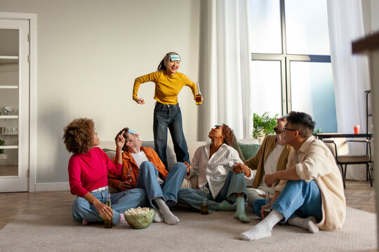 multiracial group of young student friends playing charades together and drinking beer at party at home
