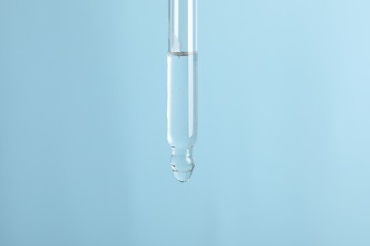 Dripping cosmetic serum from pipette on light blue background