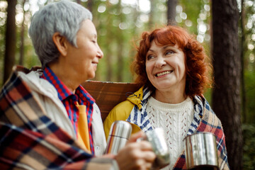 Two elderly women friends walk in the forest, pour coffee from a thermos, have a great time together