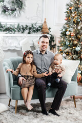 Dad spends time with his little children in front of a tree with Christmas decorations. Happy father his son and daughter playing together. Dad hugs his happy children while sitting on the sofa