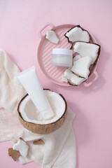 The cream jar is placed on a pink ceramic plate with fresh coconut while the cleanser tube is placed in half a coconut on a pink background. Blank labels for branding.