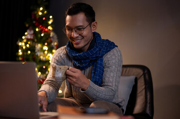 A happy Asian man is sipping coffee and using his laptop in a living room on a Christmas night.