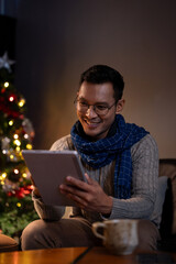 A happy Asian man is using his digital tablet while sitting at a table beside a Christmas tree.