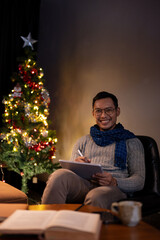 A happy Asian man in a warm cosy clothes is using his tablet while sitting beside a Christmas tree.