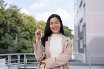 Fototapeta premium A confident Asian businesswoman is pointing her finger up while standing outdoors.