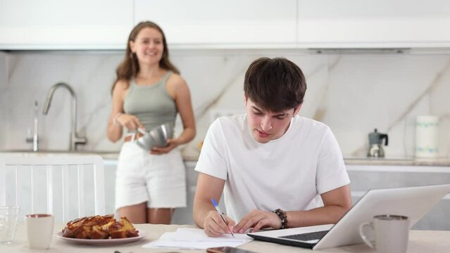 Young woman smiling and talking to young guy working at laptop in kitchen