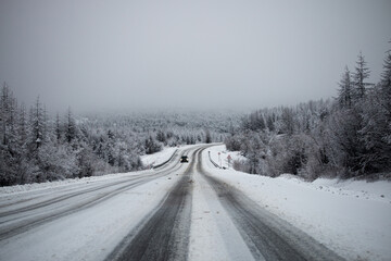 Photo of winter road during the snowfall in Magadan, Russia. Snow showers on trees and hills. Fog and haze, low visibility due to snowstorm. Extreme weather conditions. Car approaching in the distance