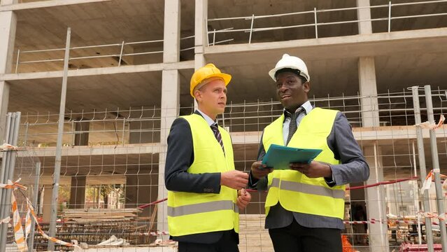 African-american and Eropean engineers standing on construction site and talking about new project. One man holding folder paper with documentation, another one pointing with finger.