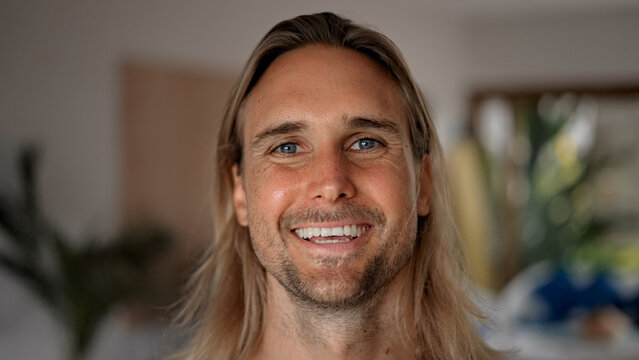 Happy male person face home portrait. Blue eyes looking at camera close up. Joyful bristle surfer man long hair. Smiling shy guy. Kind casual people. Young adult hippie inside house. Hipster hairstyle