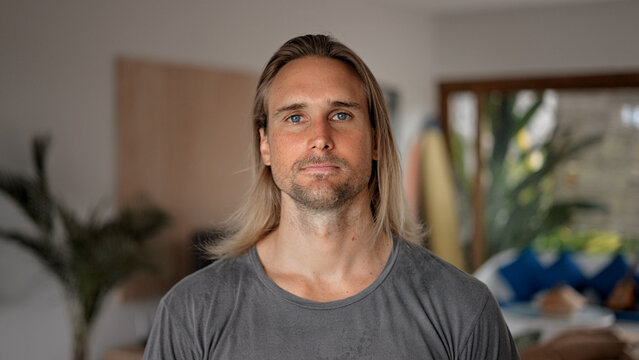 Happy male person face home portrait. Blue eyes looking at camera close up. Joyful bristle surfer man long hair. Smiling shy guy. Kind casual people. Young adult hippie inside house. Hipster hairstyle - Powered by Adobe
