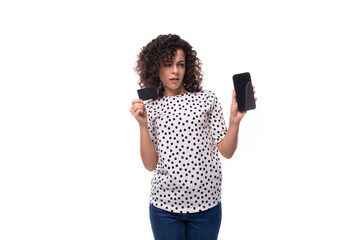 a slender young stylish woman with a curly haircut dressed in a blouse with polka dots holds a plastic card mockup and a smartphone