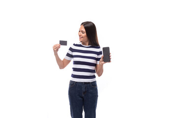 young well-groomed brunette woman with straight hair dressed in a striped t-shirt holds a plastic credit card and smartphone