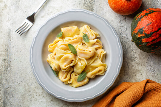Top view of plate with italian cappellacci, tortelloni, tortelli or ravioli stuffed with pumpkin and parmesan cheese, nutmeg and sage leaves. Whole pumkins.  Light grey texture table.