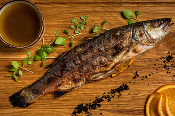 Top view close-up of whole grilled sea bass fish on a wooden board with orange, microgreens, sauce